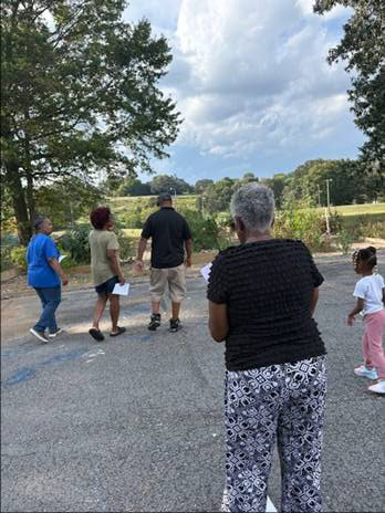 A diverse group of people gathers outdoors at a community garden in Knox County, engaging in educational activities and socializing amidst lush greenery and clear skies.