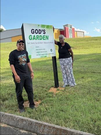 Young woman and young man standing near a large God's Garden sign in a community garden setting.