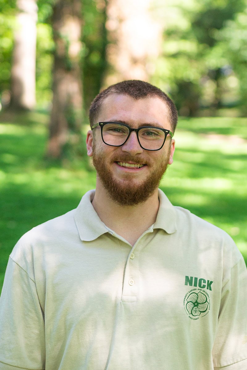 Bright young man with glasses and a beard smiling outdoors at Knox County Community Garden, wearing a beige polo with the "NICK" logo, representing Knox County's urban gardening program, surrounded by lush green trees.