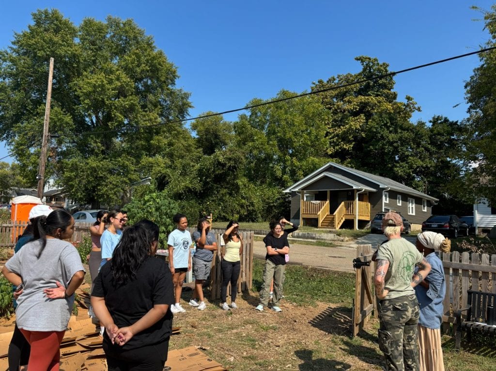 Volunteer group gathered outdoors at Knox County Community Garden on a sunny day, engaging in gardening activities and community building among lush green trees.