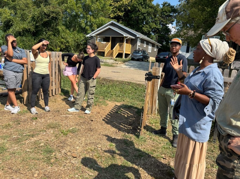 People gathered outdoors at a community garden, listening to a woman speak and share information, with a small wooden fence and residential houses in the background.