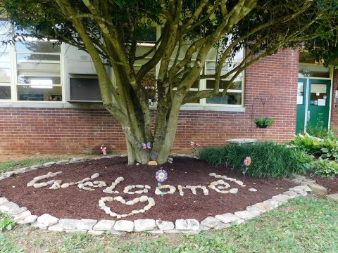 Community garden with a large tree, flower bed spelling "Welcome" with a heart, surrounded by rocks, located in front of Inskip Elementary School.