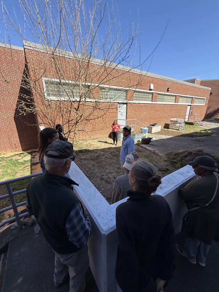 Group of community garden members gathered outdoors near a brick building during a meeting, discussing gardening activities and community projects.