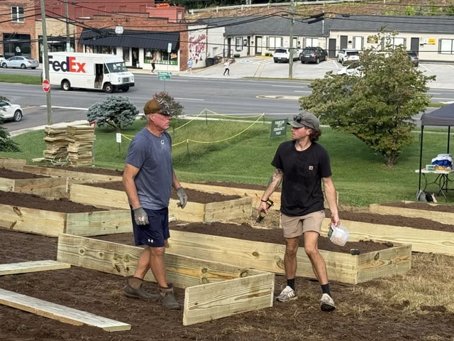 Two men working together on a community garden project with wooden raised beds and a city street in the background.