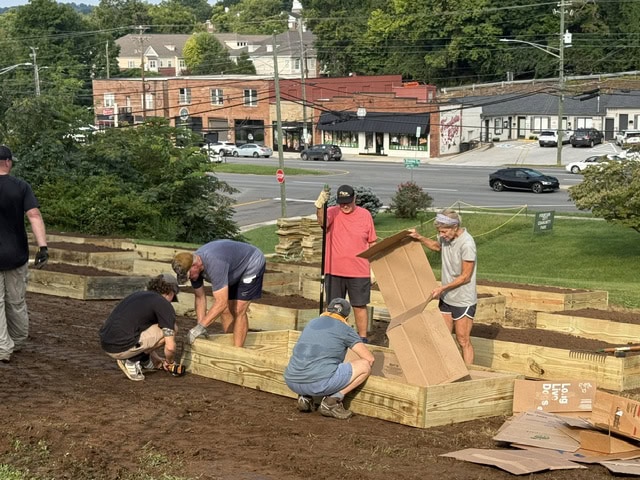 Group of people assembling wooden garden beds at a community garden site with urban background and trees.