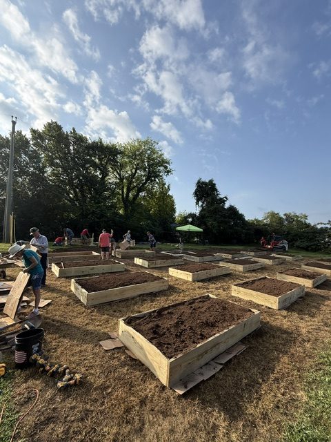 People working on building raised garden beds in a community garden under a blue sky with clouds, surrounded by trees.