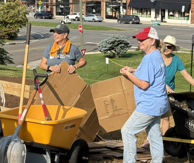 Women planting and preparing soil for a community garden, with cardboard and gardening tools, in an urban setting.