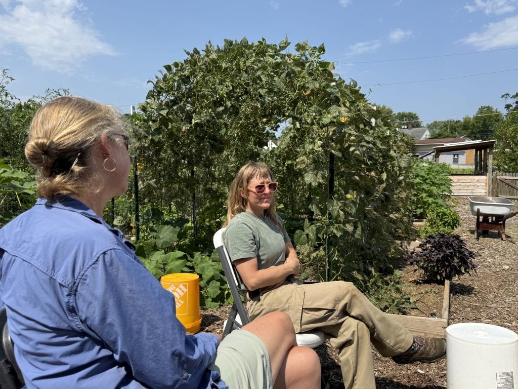 Two women sitting and talking in a community garden with lush greenery and a clear blue sky.