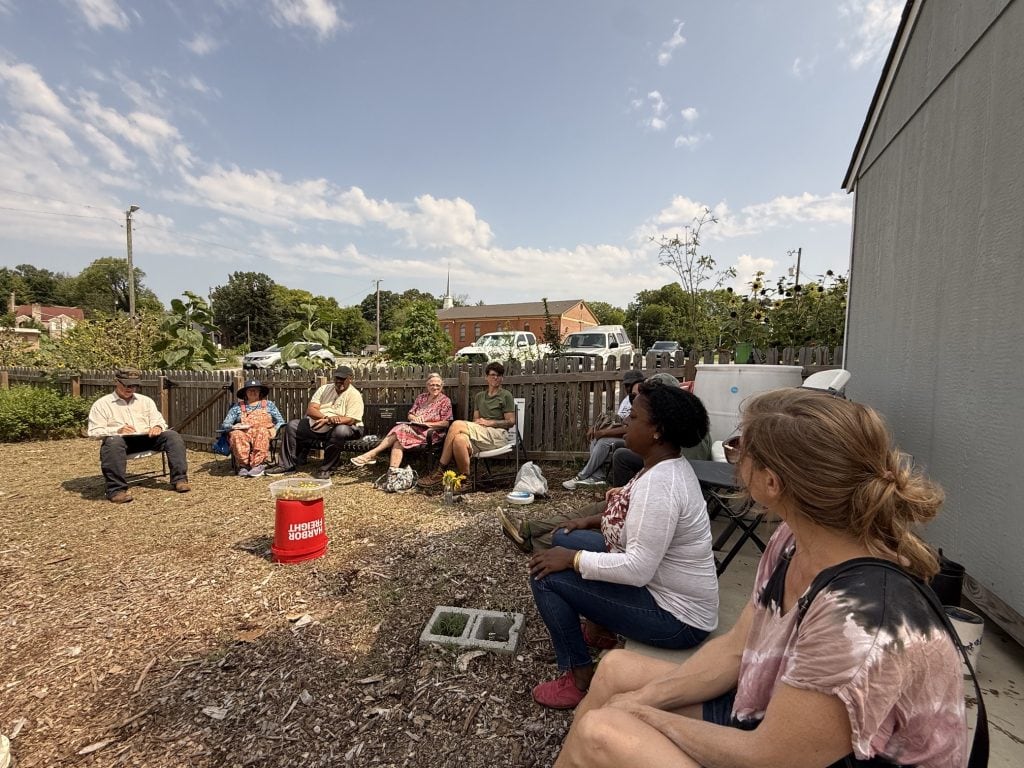Community members gather outdoors at Knox County Community Garden for a meeting during a sunny day.