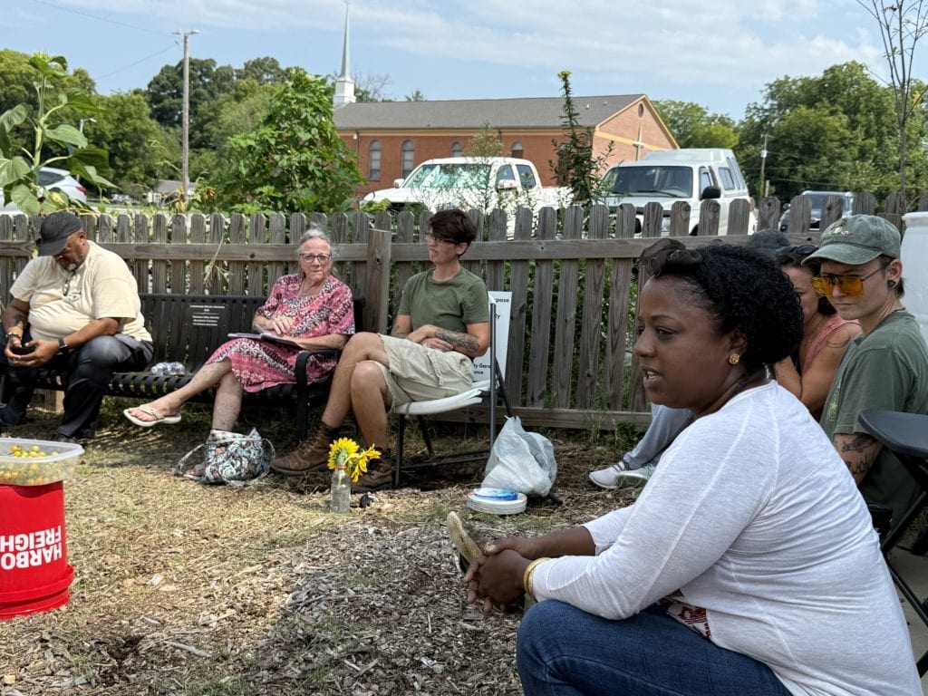 Community members gathered outdoors at Knox County Community Garden for a meeting, sitting on chairs and benches, with a wooden fence and trees in the background.