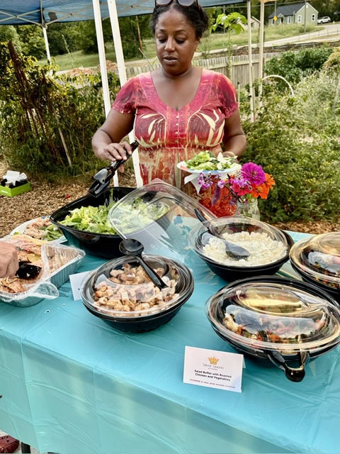 Woman serving food at a community garden dinner event with various dishes on a table and a garden background.