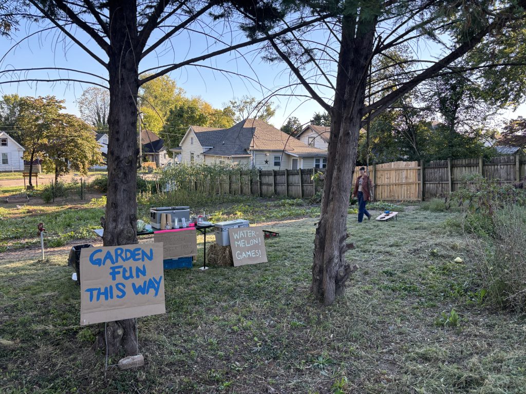Community garden with signs for garden fun and watermelon games, featuring trees, houses, and a person in the background during daytime.