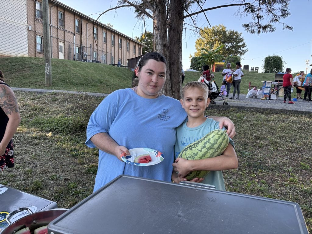 Two children at Knox County Community Garden & Growers Alliance event, one holding a watermelon, outdoors during daytime with other people in background.